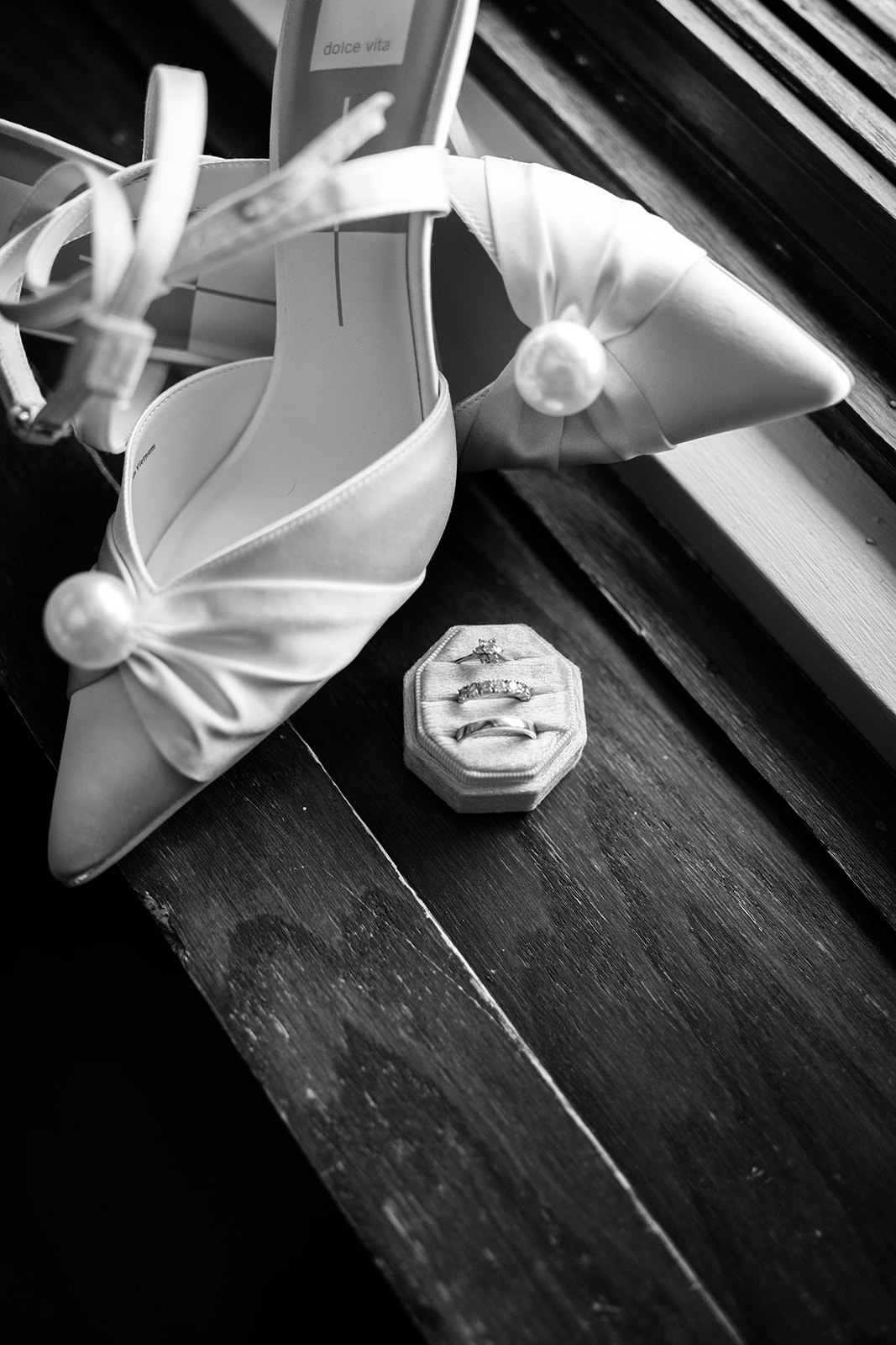 Black and white film photo of silk bridal heels with pearl accents and rings on a wooden windowsill.